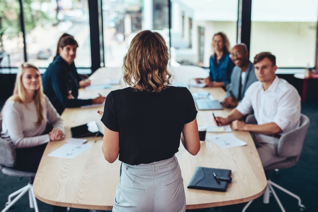 A woman is giving a presentation to a group of people sitting around a table.