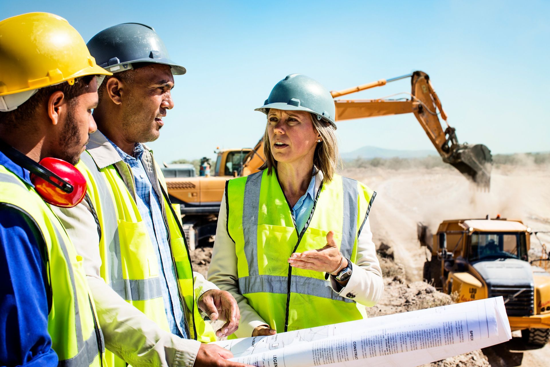Mining engineers and workers in safety gear discuss a project with heavy machinery operating in the background.