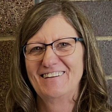 A woman wearing glasses is smiling in front of a brick wall.