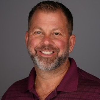 A man with a beard and a purple shirt is smiling for the camera.