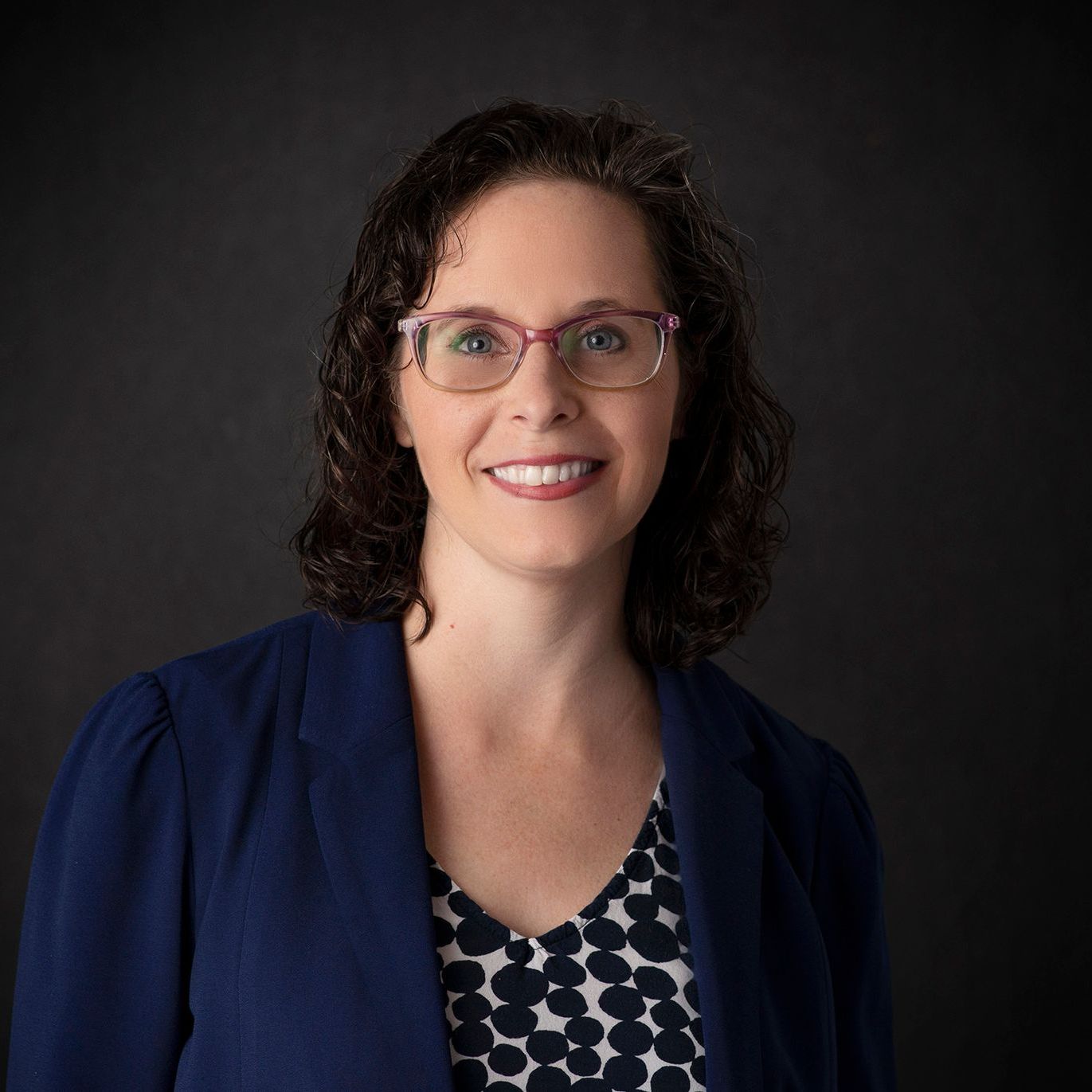 A woman wearing glasses and a blue jacket is smiling for the camera.