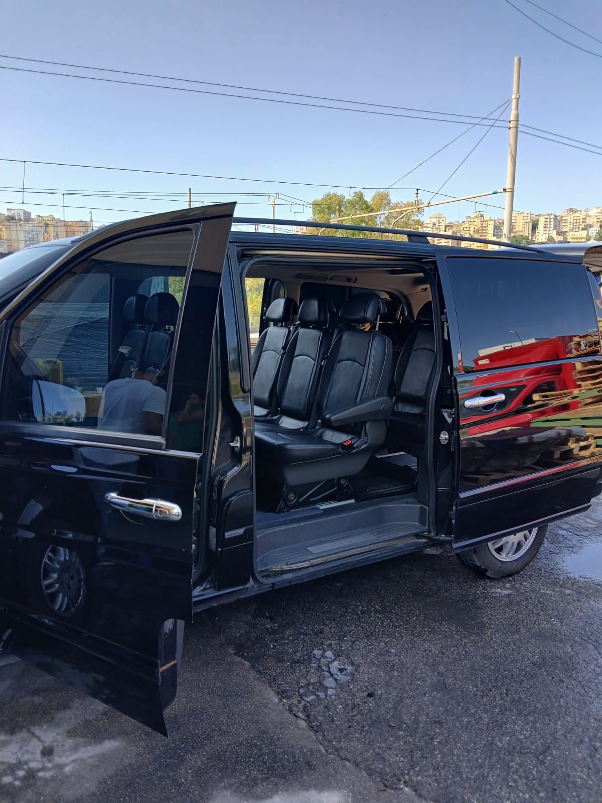 Viaggiatori con bagagli salgono a bordo di un furgone Mercedes nero fuori da un terminal aeroportuale.
