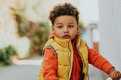 Child in a yellow vest and orange sleeves standing outdoors, looking at the camera.