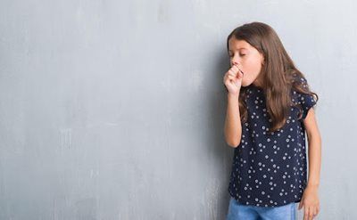 Person in a navy polka-dot shirt coughing against a gray wall