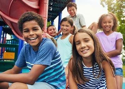 Children smiling on a playground slide, with a red tunnel slide and colorful play area behind them