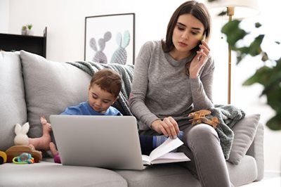 Woman talking on phone while child uses a laptop on a gray couch in a living room.