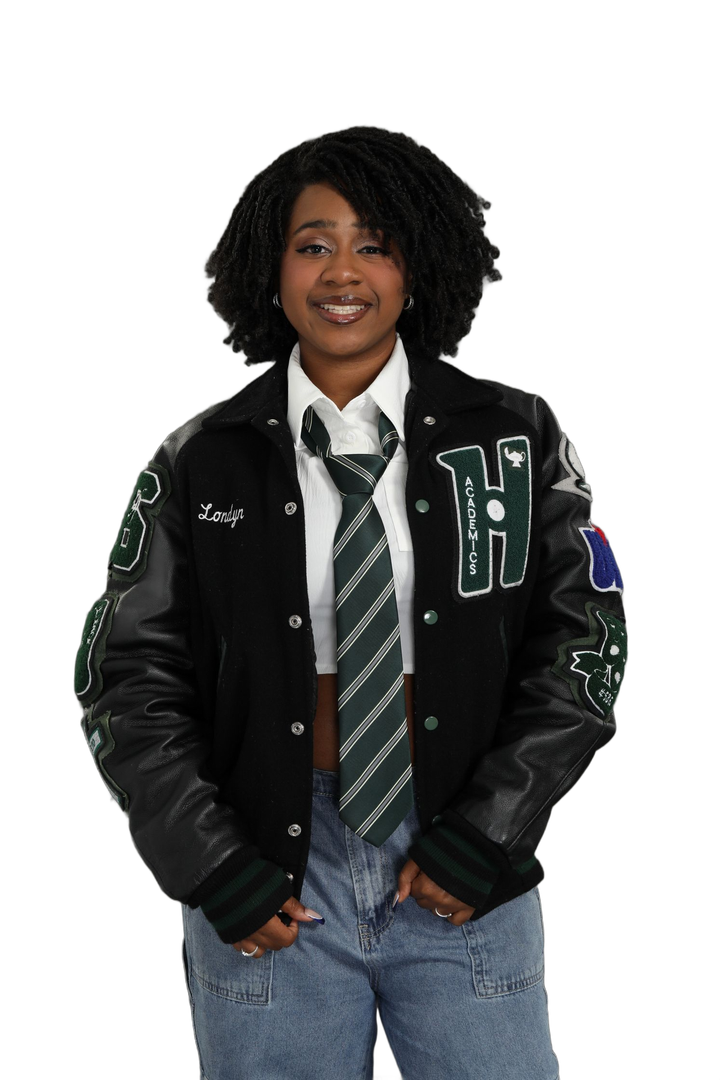 Person posing against a white background in a black letterman jacket, tie, jeans, and hands in pockets