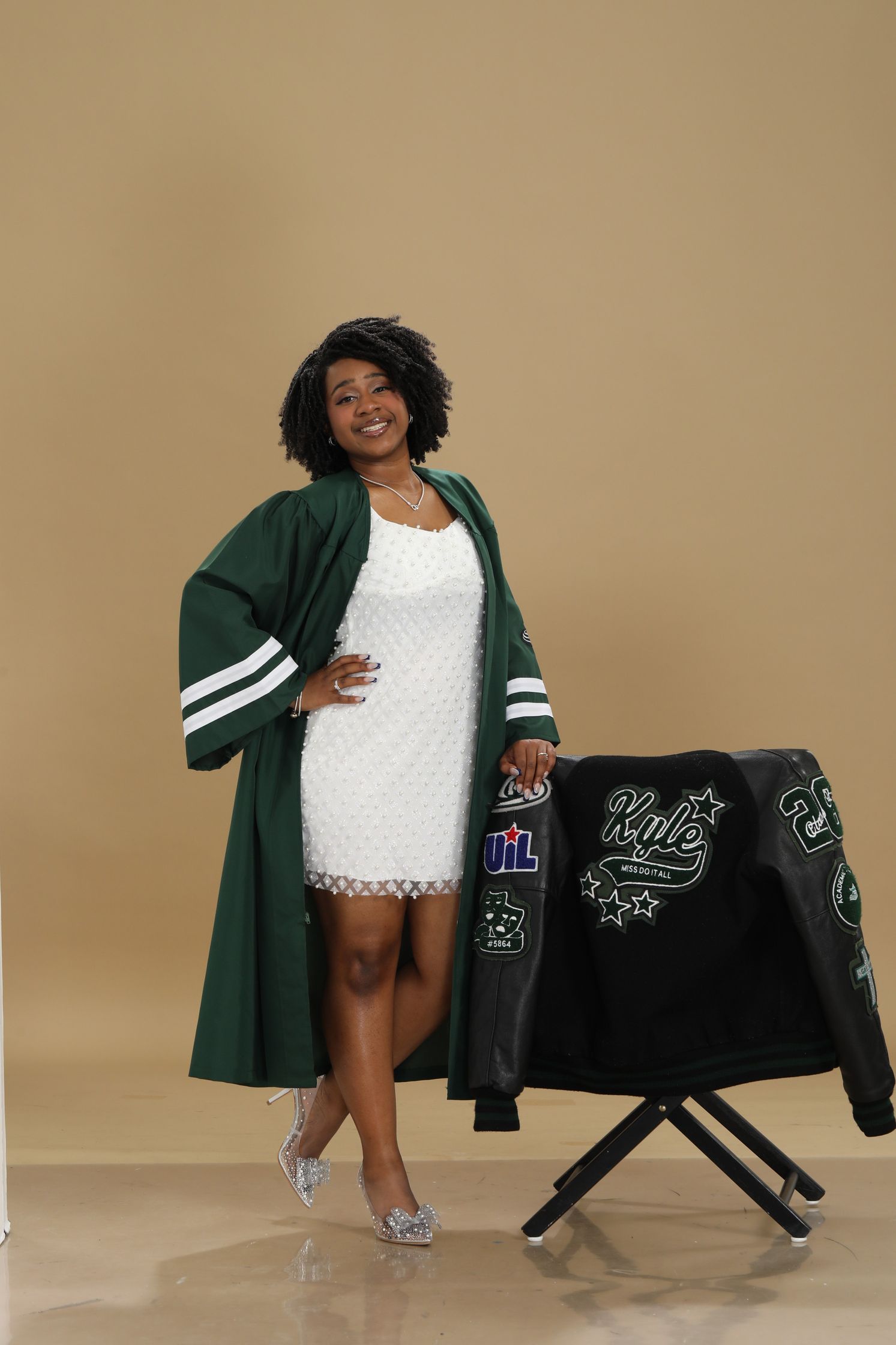 Londyn in a green graduation robe and white dress standing beside a decorated clothing display against a beige backdrop