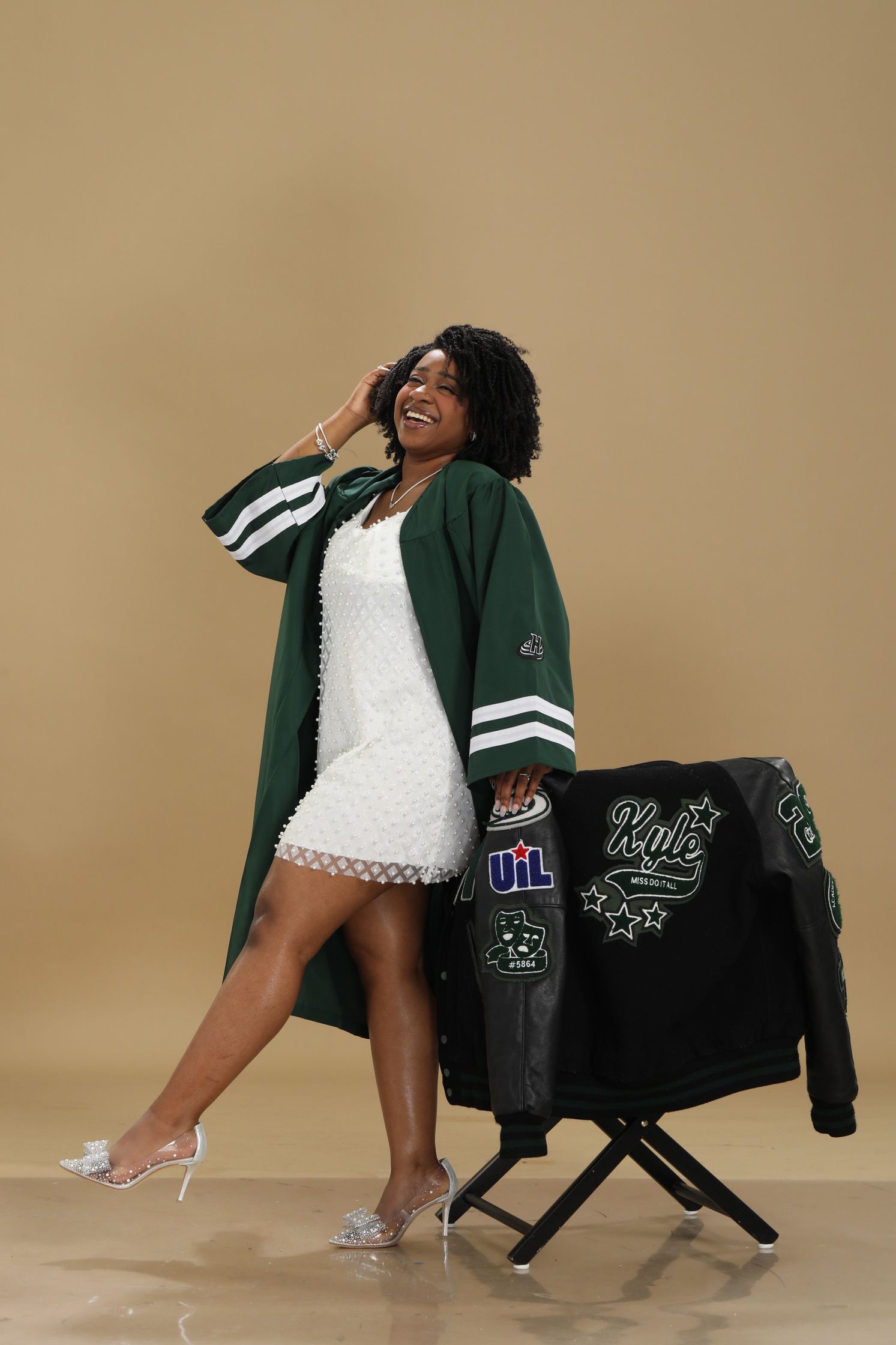 Londyn in a green graduation robe posing beside a garment rack on a beige backdrop