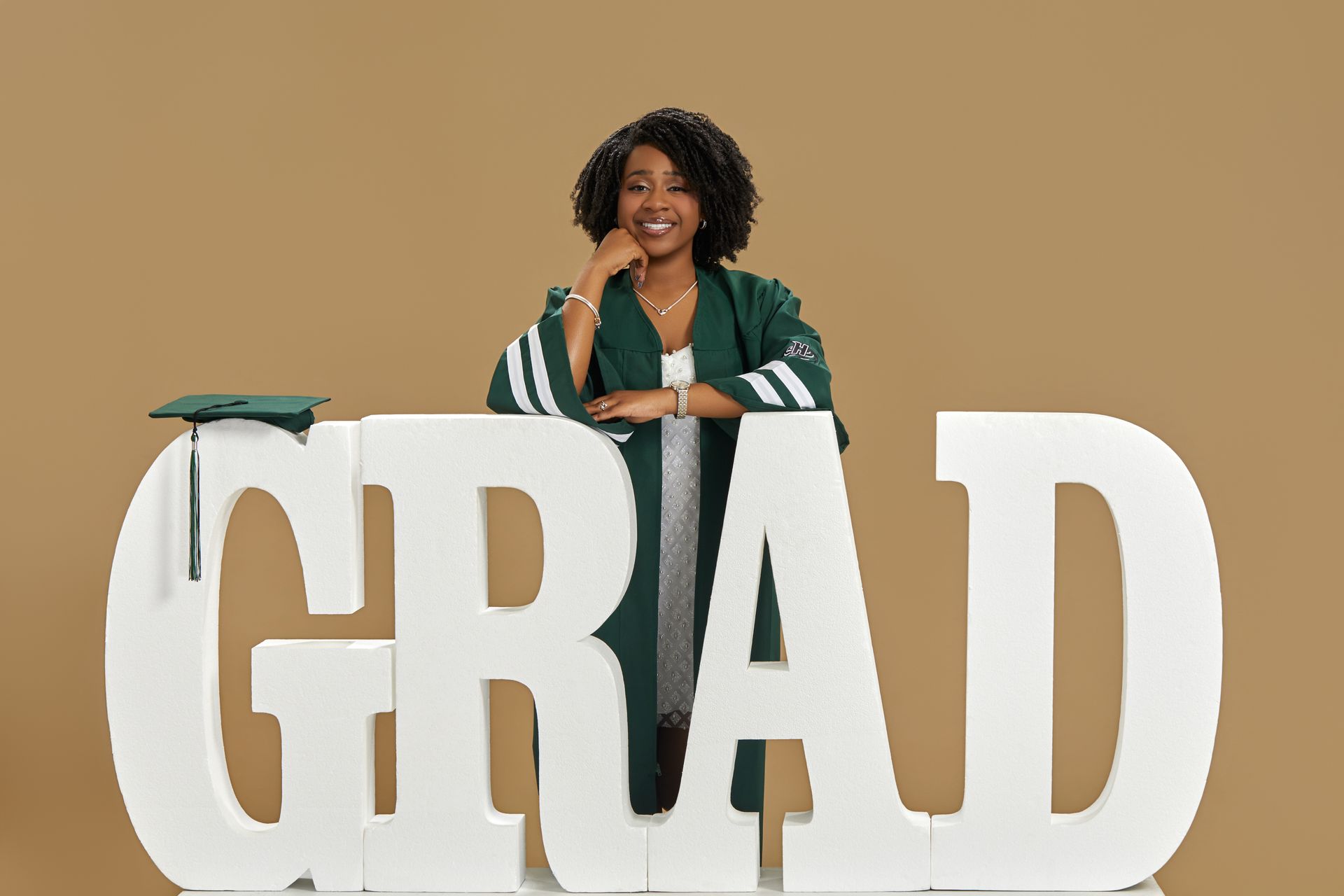 Graduate smiling behind large white “GRAD” letters against a tan background