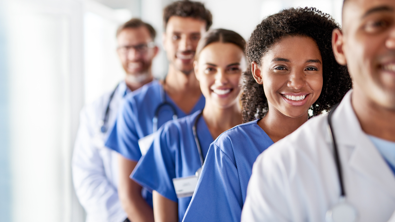 Healthcare team smiling in a row, wearing scrubs and lab coats in a bright setting.