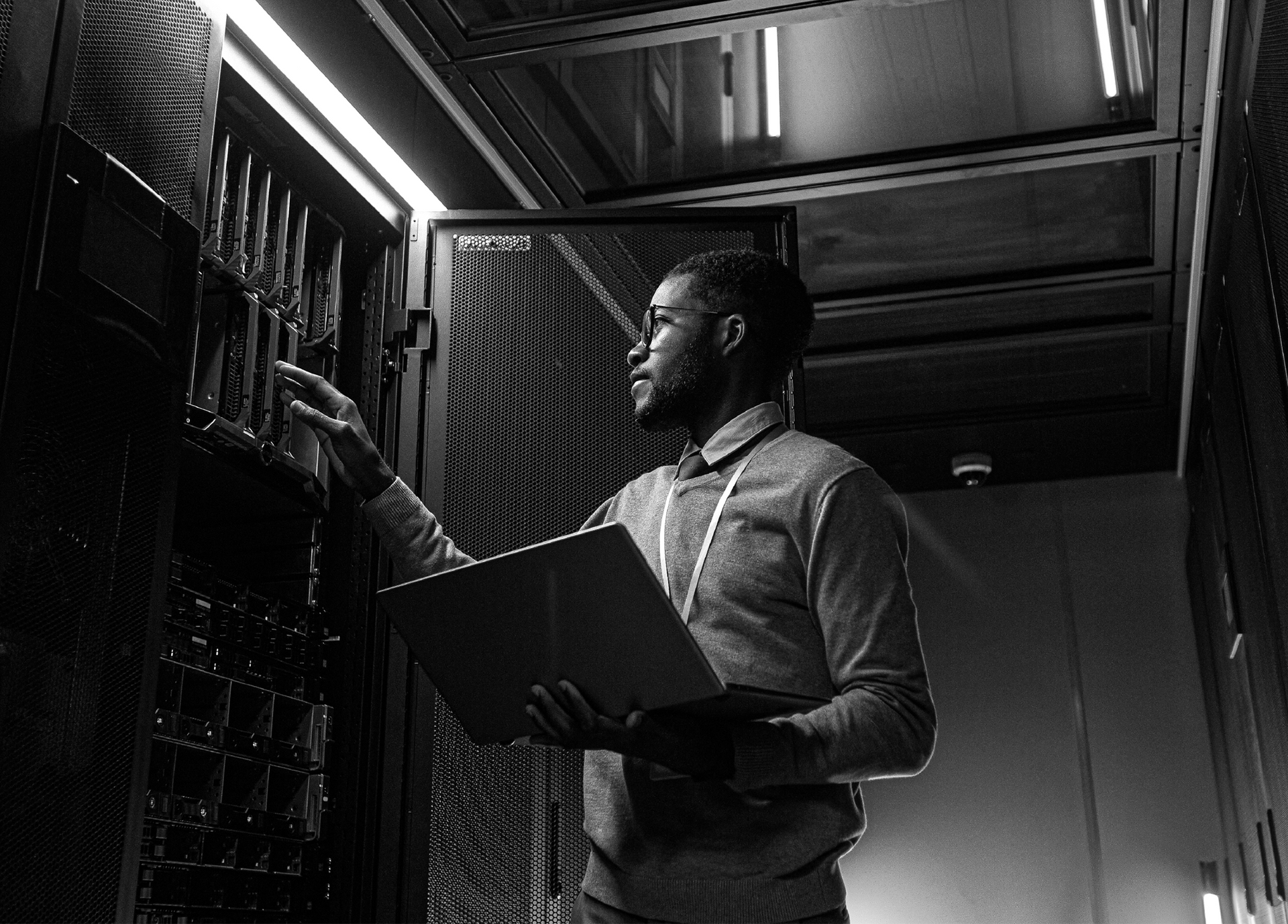 Profissional de TI monitorando servidores em data center com notebook em mãos.