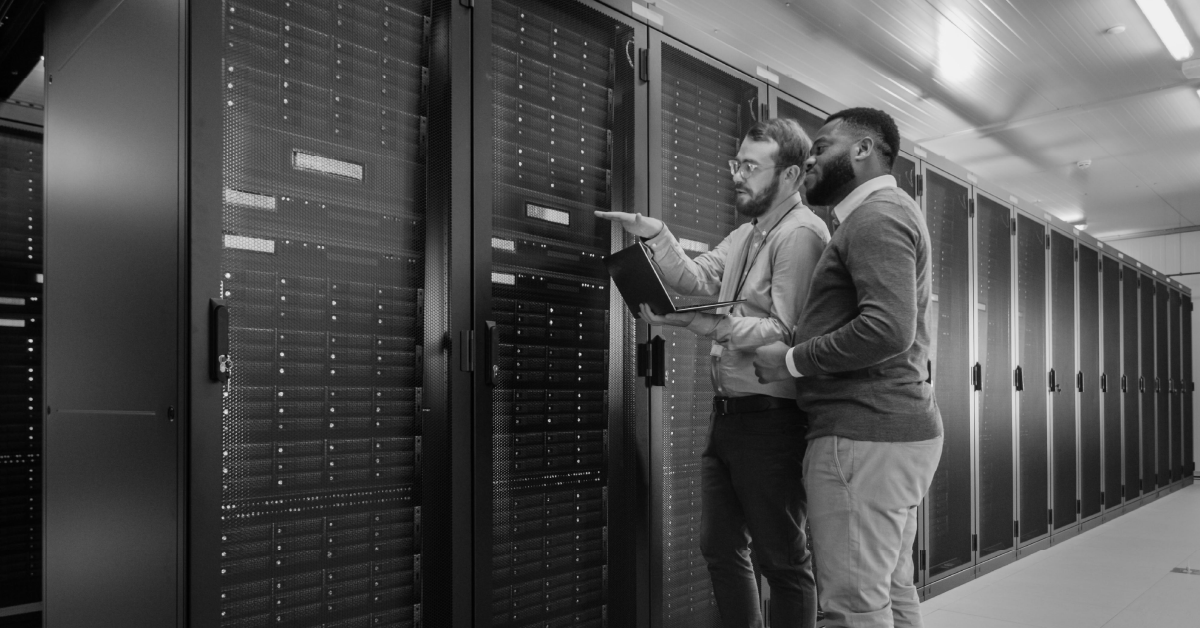 Dois profissionais em P&B analisam racks de servidores em um data center. Um com laptop aponta.