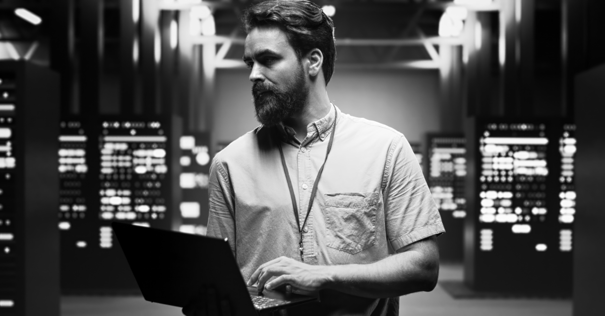 Homem com camisa casual e crachá, segurando um laptop, olha para o lado em um data center com racks.
