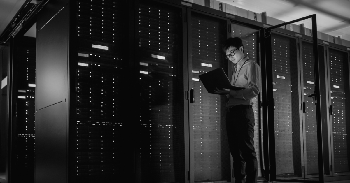 Homem com óculos e laptop, parado em corredor data center, observando os servidores com luzes.