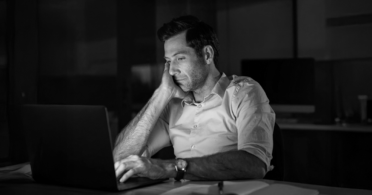 Profissional pensativo em escala de cinza, trabalhando no laptop em ambiente escuro.
