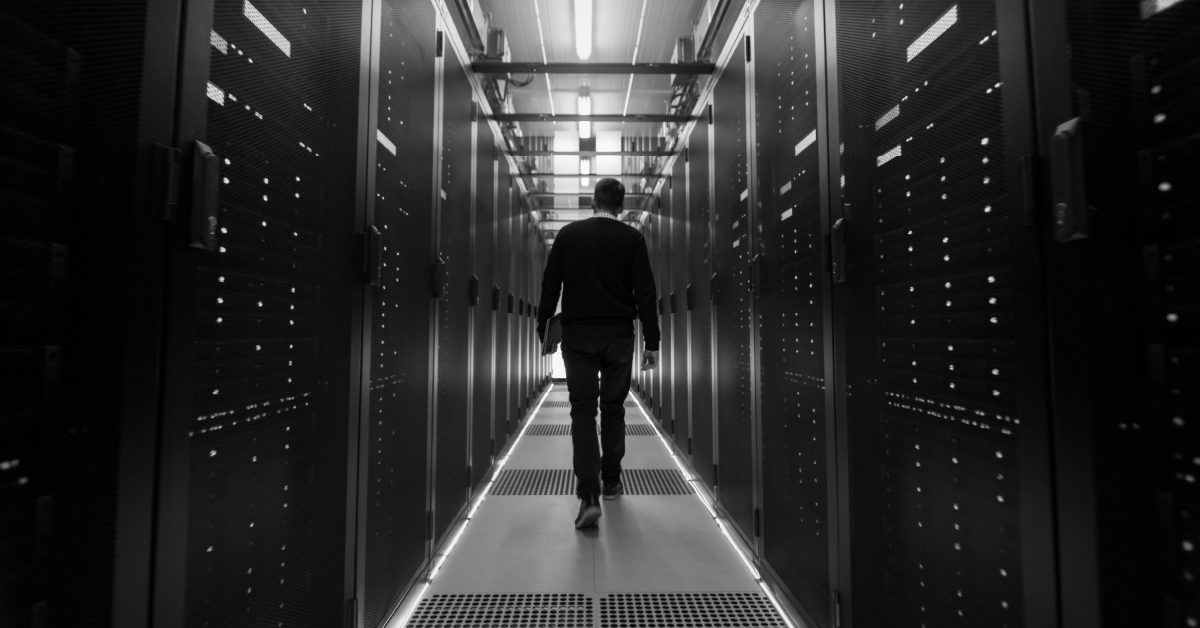 Homem de costas caminhando em corredor data center, fileiras de racks de servidores com luzes acesas