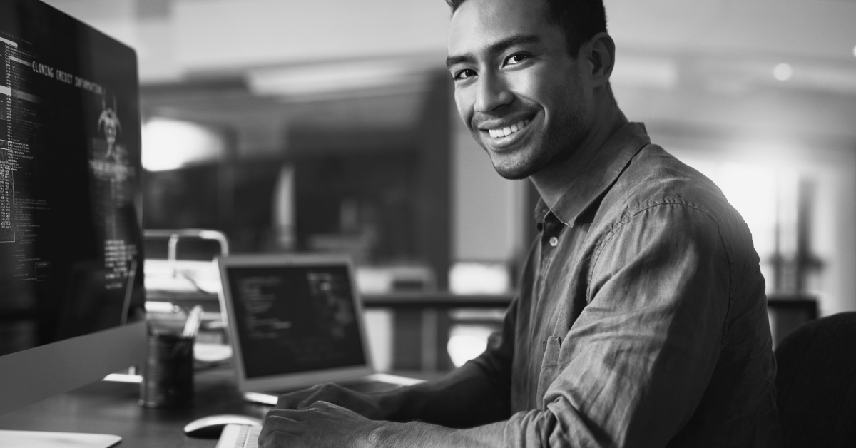 Homem trabalhando em um laptop com código na tela ao fundo e sorrindo. Imagem em preto e branco.