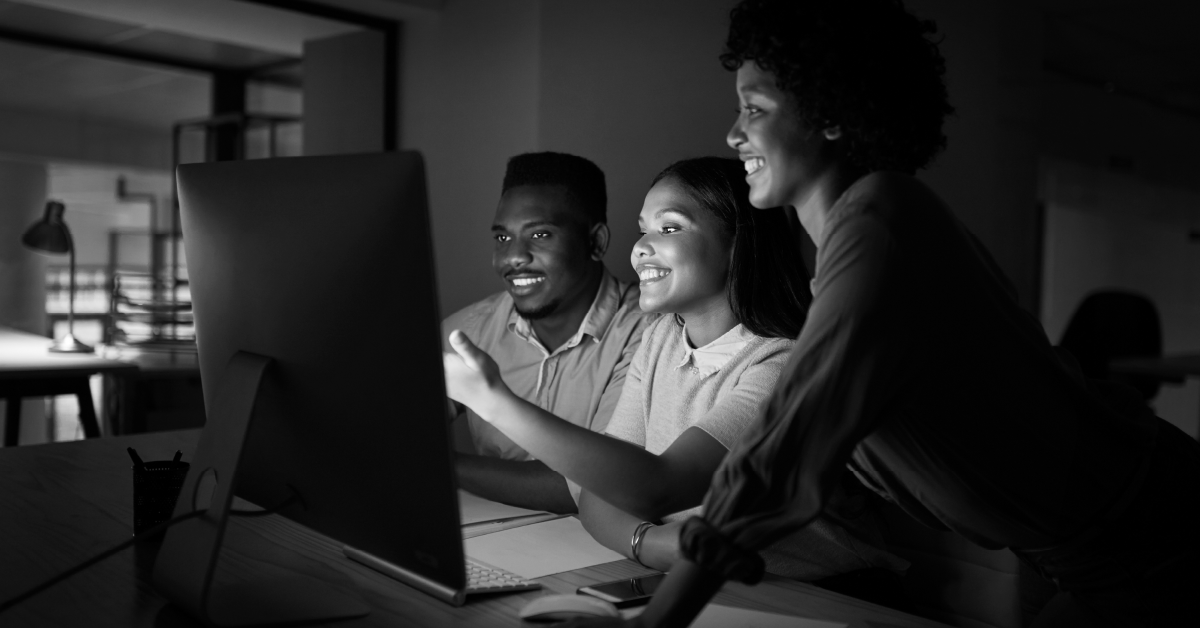 Três pessoas sorrindo e olhando juntas para a tela de um computador. Imagem em preto e branco.