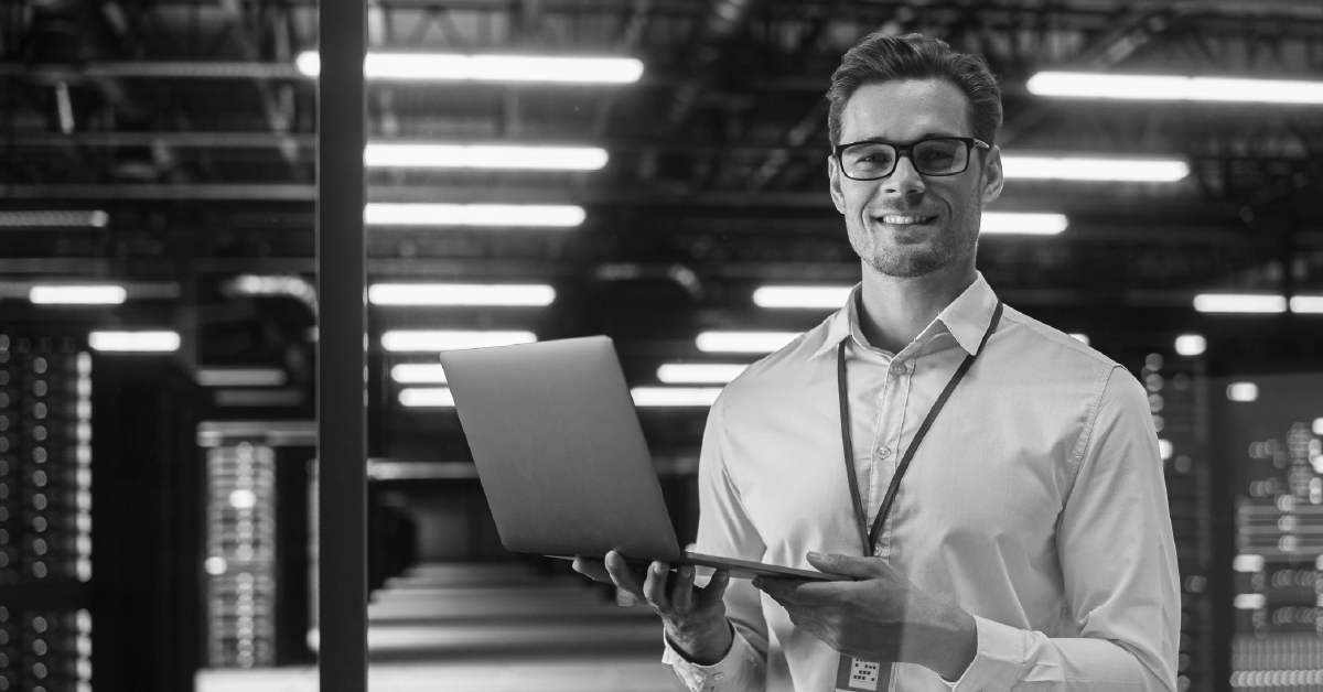 Homem sorrindo segurando laptop. Imagem em preto e branco.