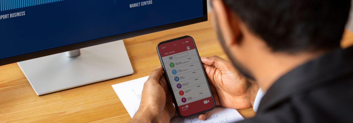 Profissional analisando e-mails no celular enquanto monitora dados em tela de computador