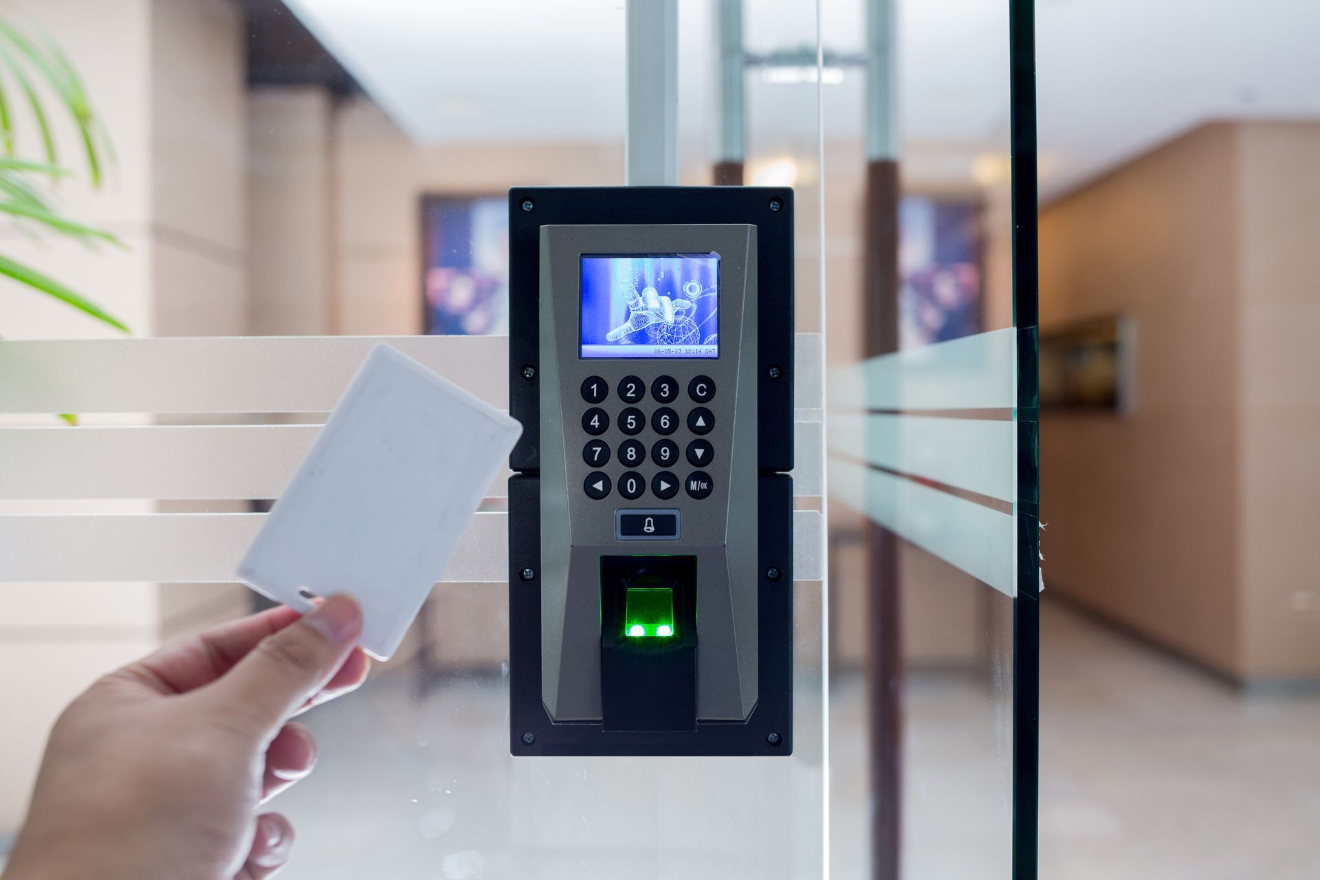 Hand holding a white card near a black access control system with a keypad, fingerprint reader, and screen on a glass door.