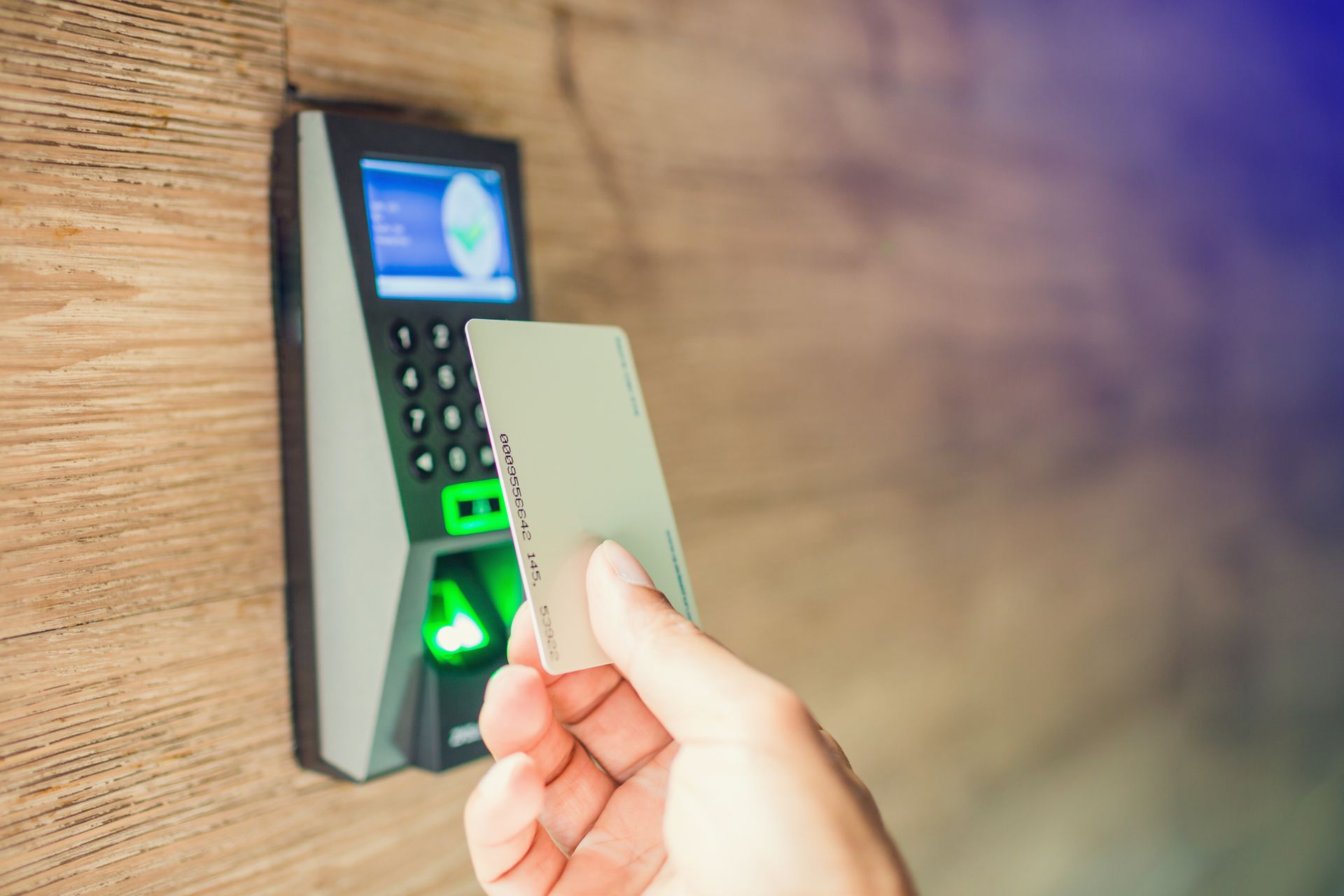 Hand holding a white card to a security access reader on a wooden wall.