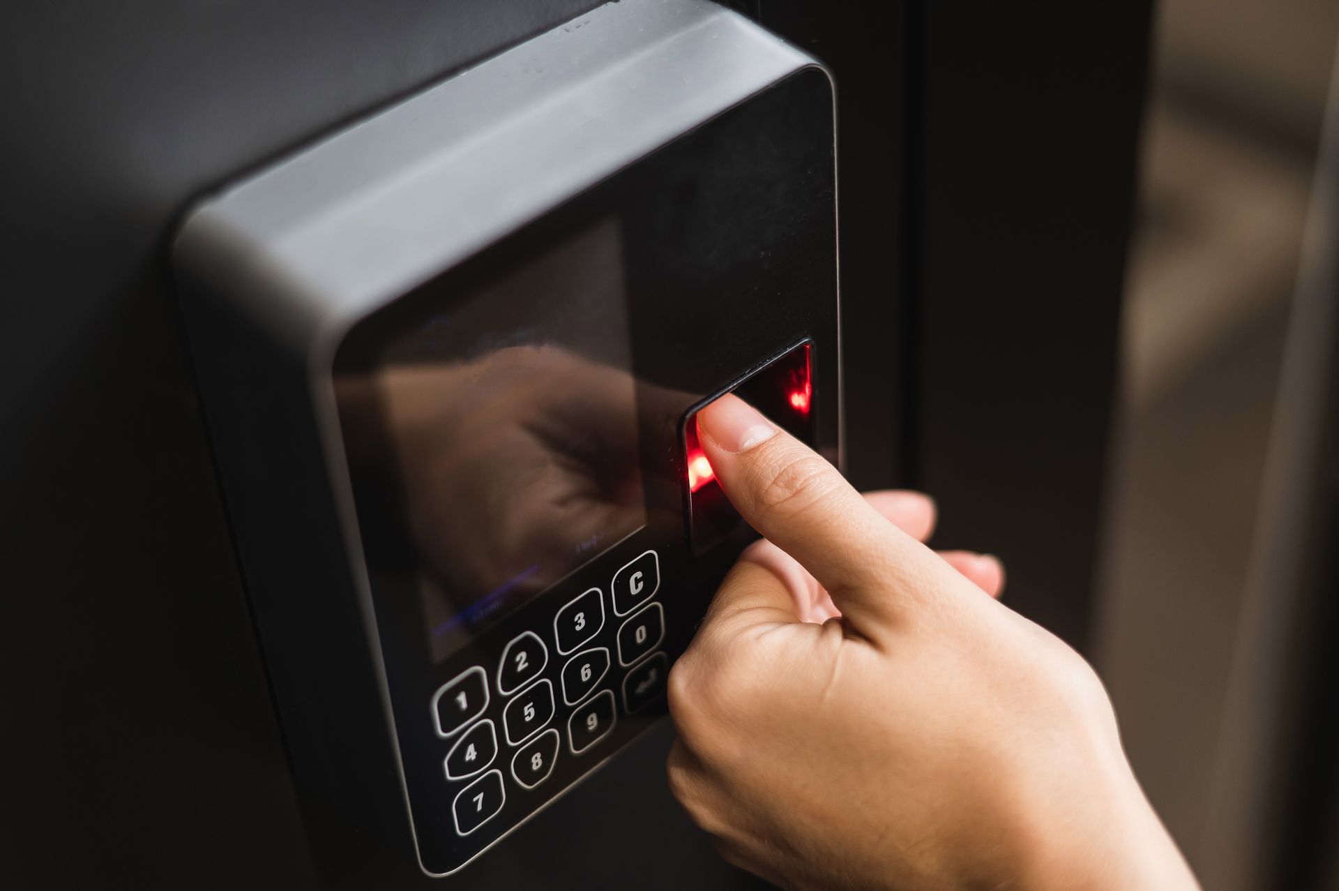 Hand pressing a fingerprint scanner on a black keypad with a red light.