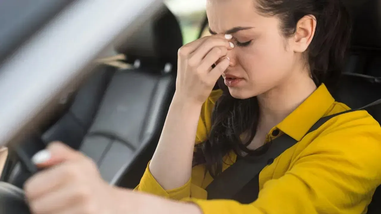 Woman in a car, appears stressed, holding her nose with eyes closed. Yellow shirt, black interior.