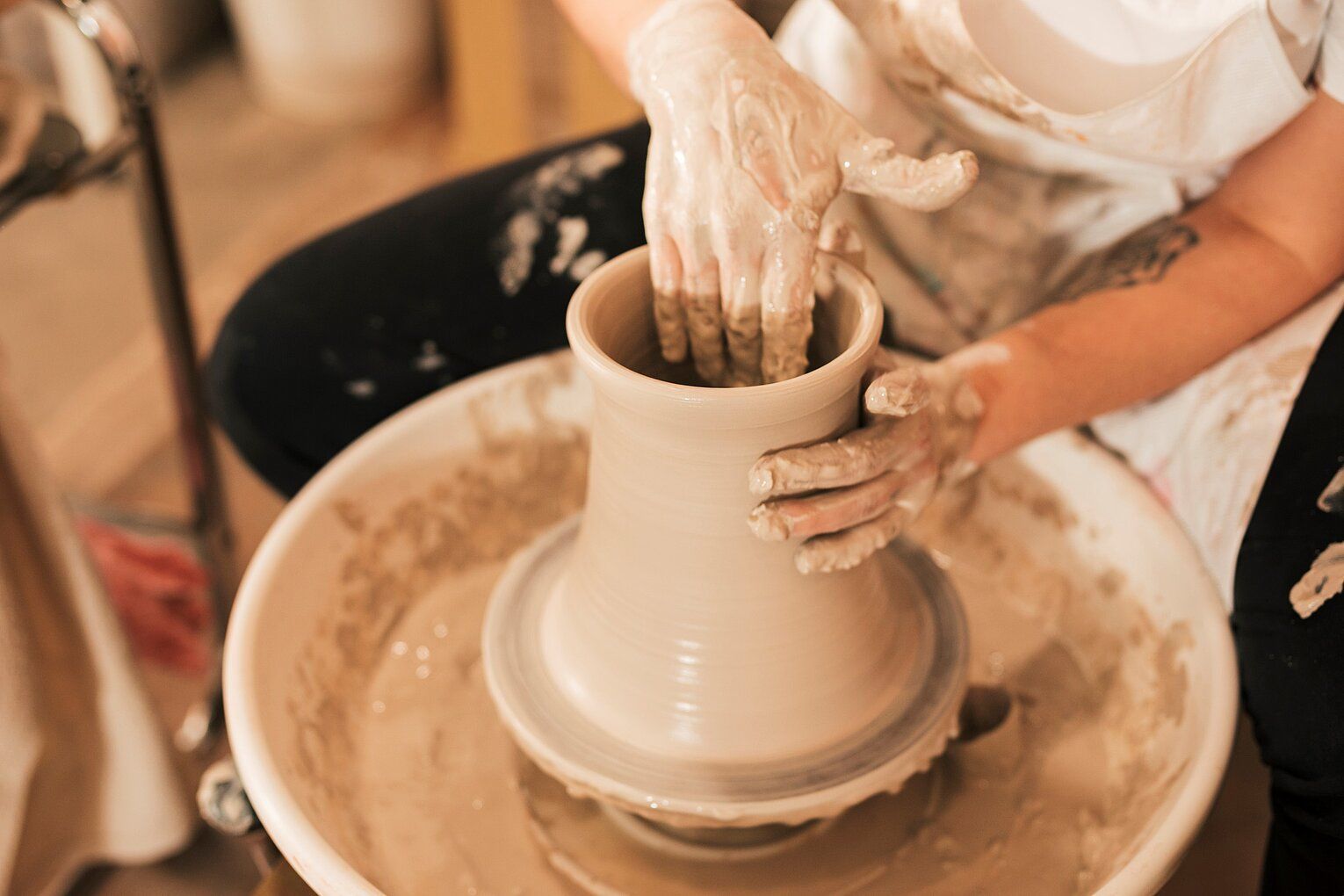 Person shaping clay vase on a pottery wheel; hands covered in clay, light brown, indoors.