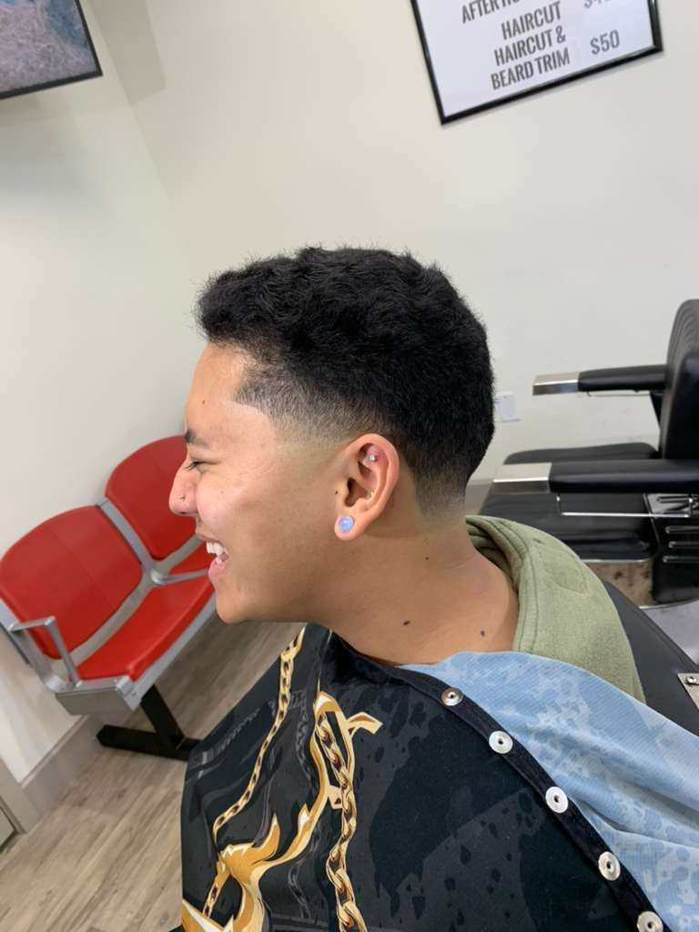Person smiling, with a fade haircut, in a barbershop, wearing a barber's cape.