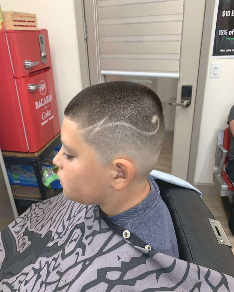 Boy with a faded haircut and decorative design on his head, sitting in a barber chair.
