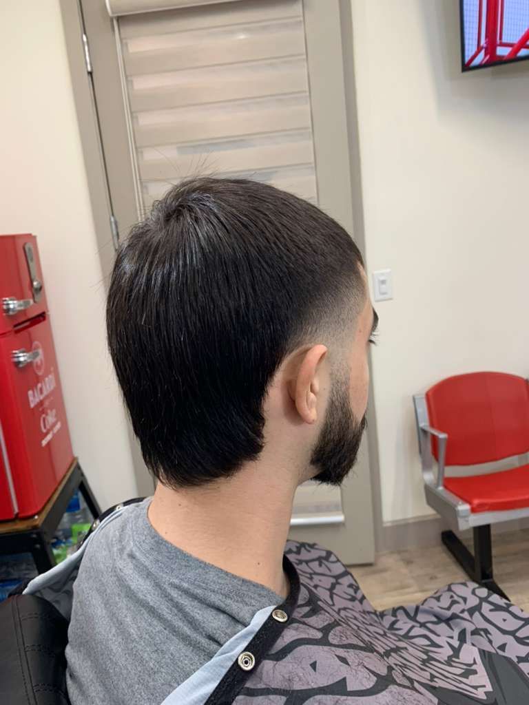 Man with short, dark hair and beard, wearing a gray shirt, getting a haircut at a salon.