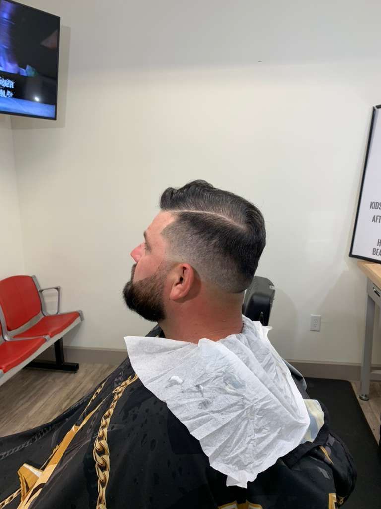 Man in barber chair, getting a fade haircut. He has a beard, a paper bib, and a black cape.