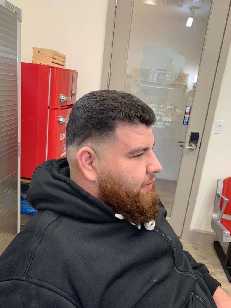 Man with dark hair and beard getting a haircut in a barbershop; faded sides, red fridge in background.