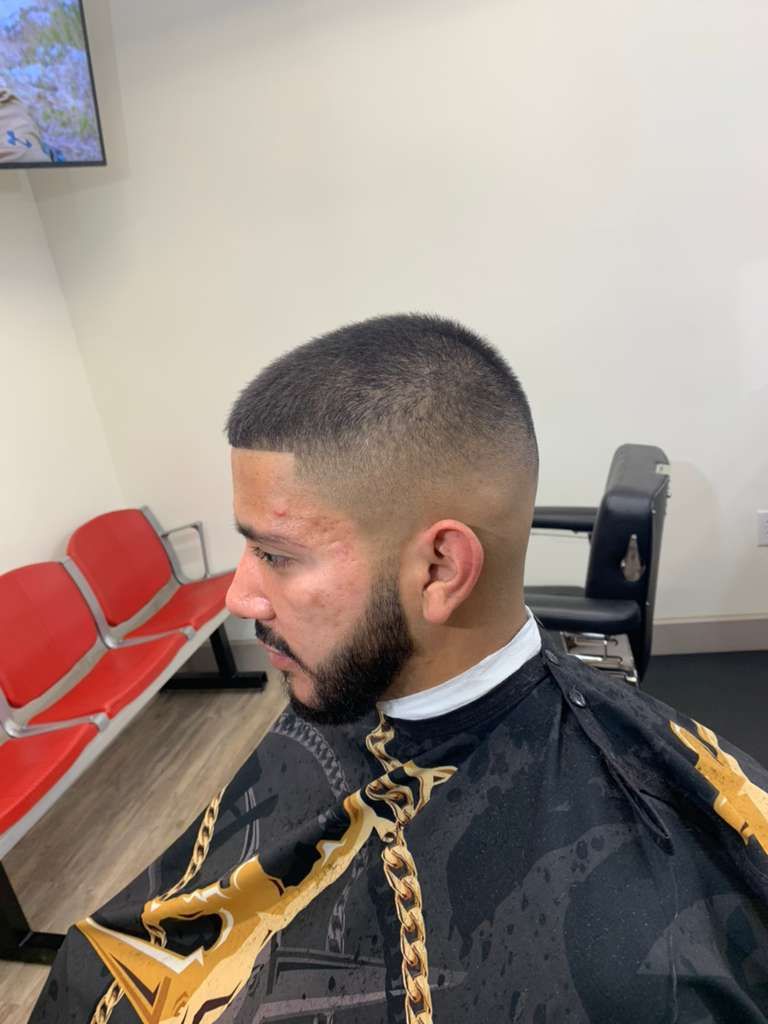 Man with a faded haircut and beard at a barbershop, wearing a cape, seated with red and black chairs in the background.