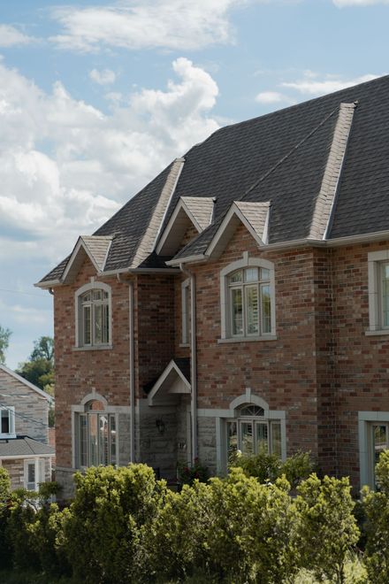Brick townhouse with dark roof and white-trimmed windows behind green shrubs under a partly cloudy sky