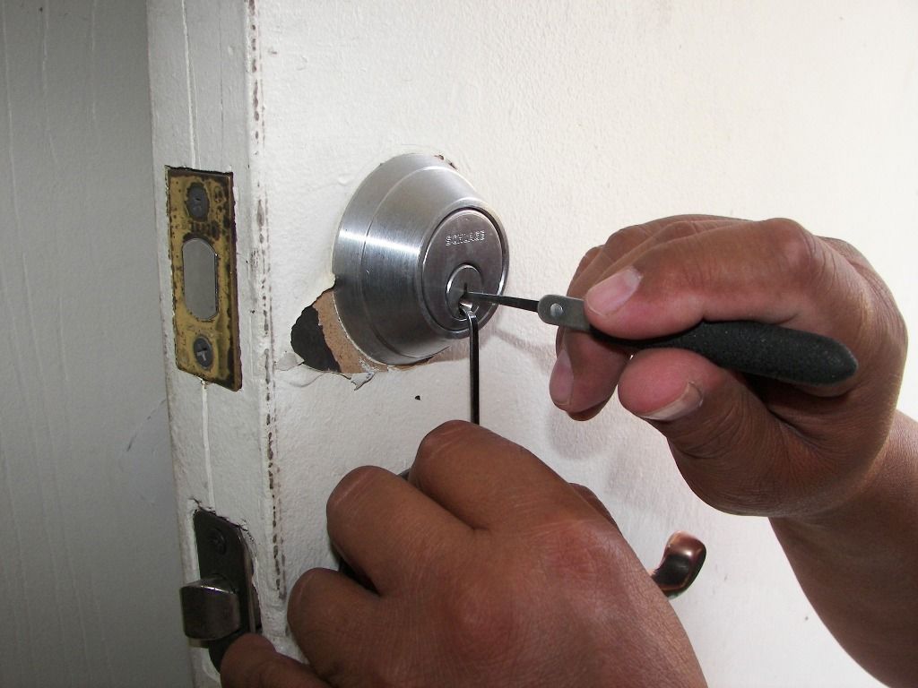 Hands picking a lock on a white door. A pick tool is in the keyhole, another in the bottom hand.