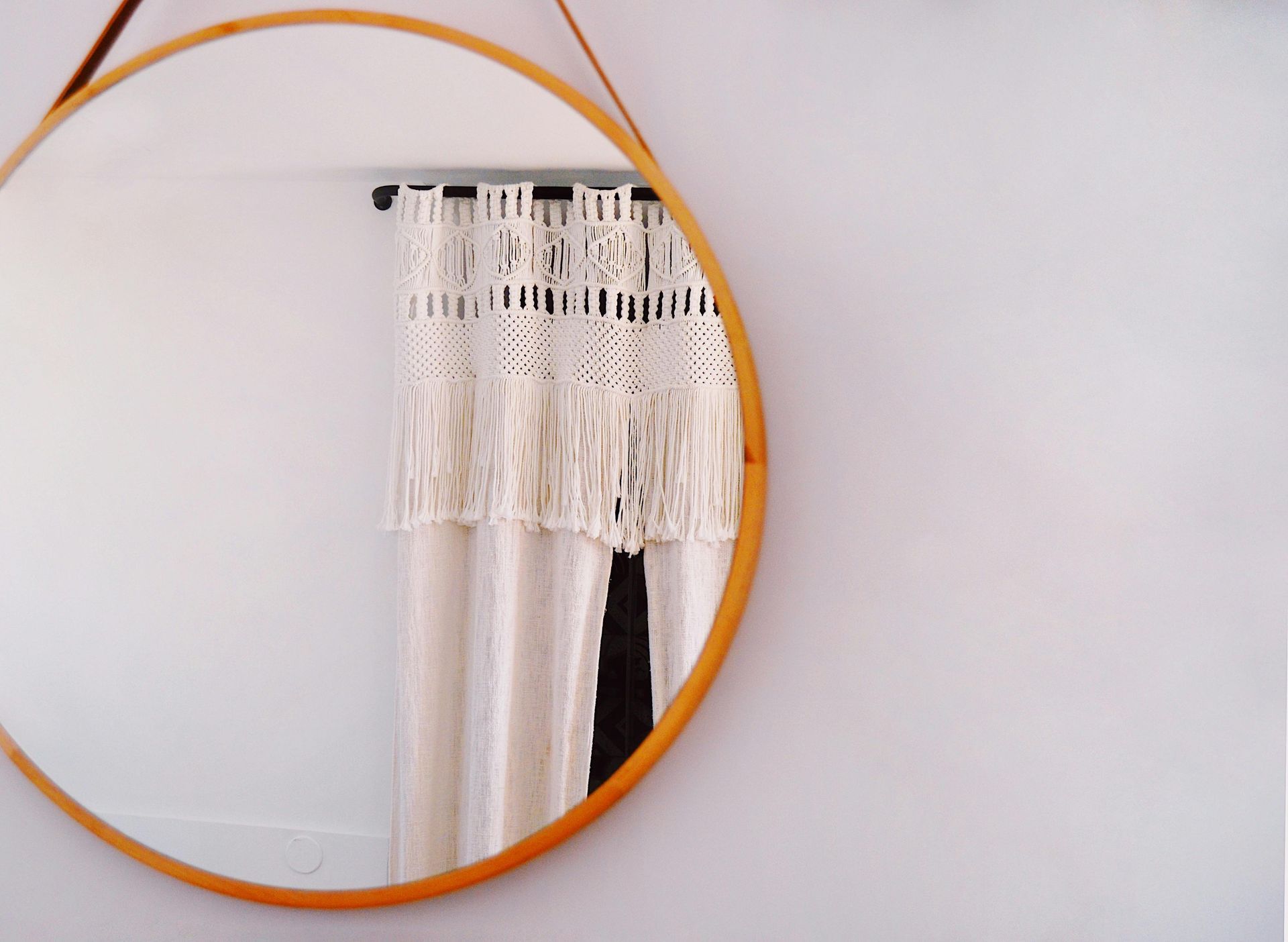 Round wooden mirror reflecting white wall and macrame curtain.