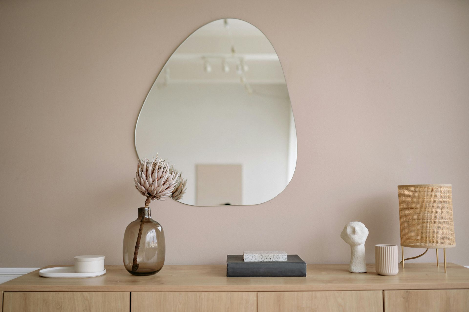 Mirror and decor on a light wood cabinet against a light brown wall; includes a lamp, vase, and sculptures.