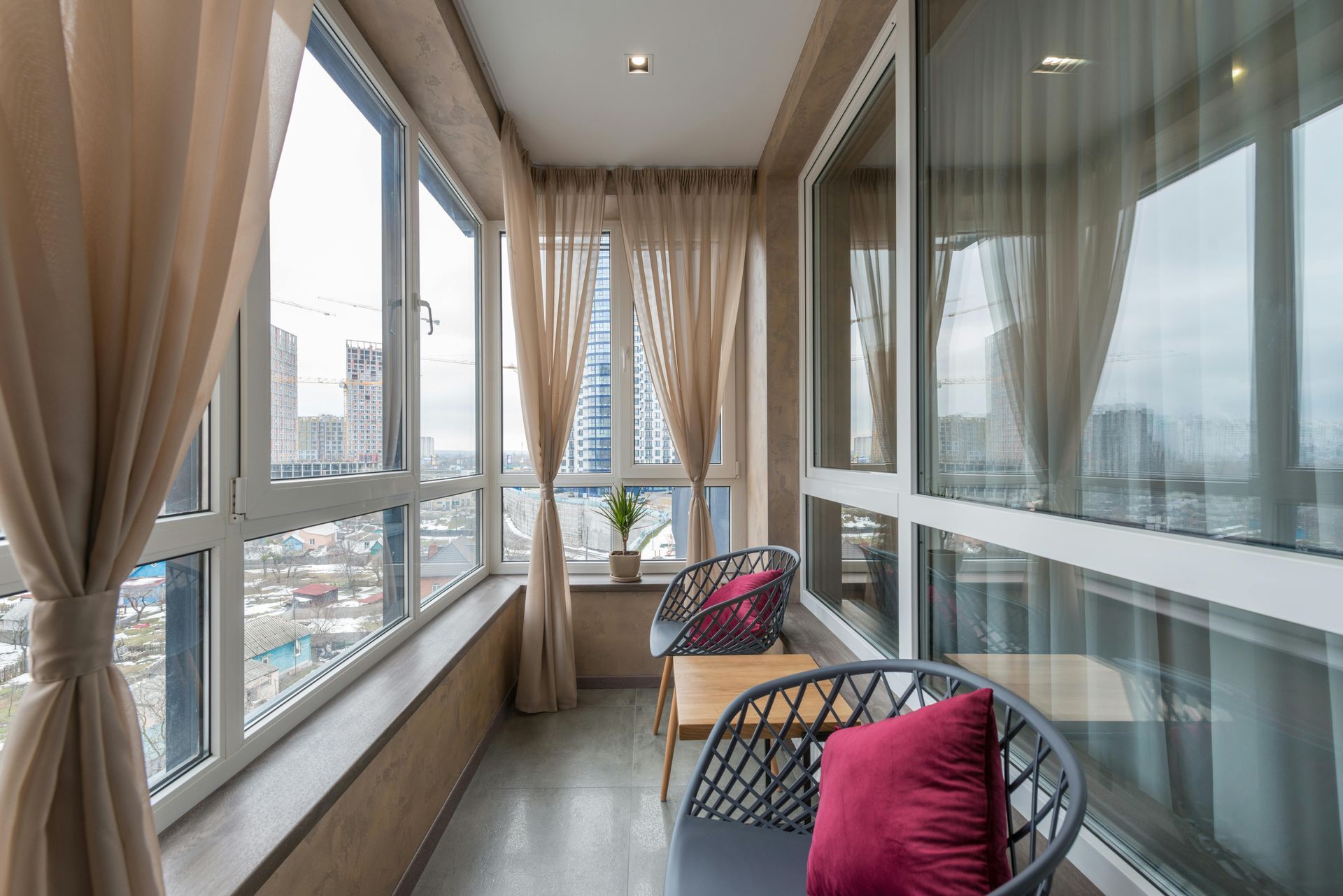 A small, modern balcony with city views. Features sheer curtains, two chairs with cushions, and a small table.