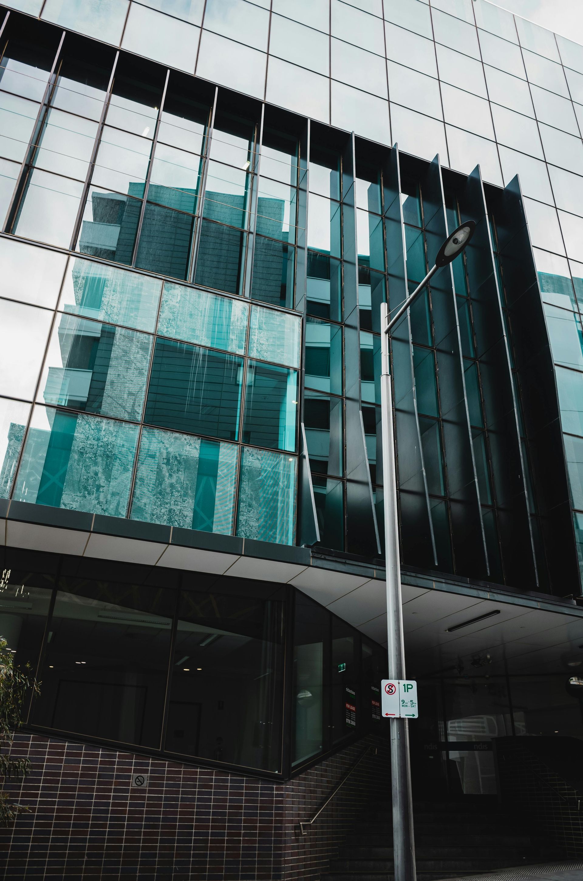 Modern glass building reflecting the sky and nearby structures, with a lamppost and steps in front.