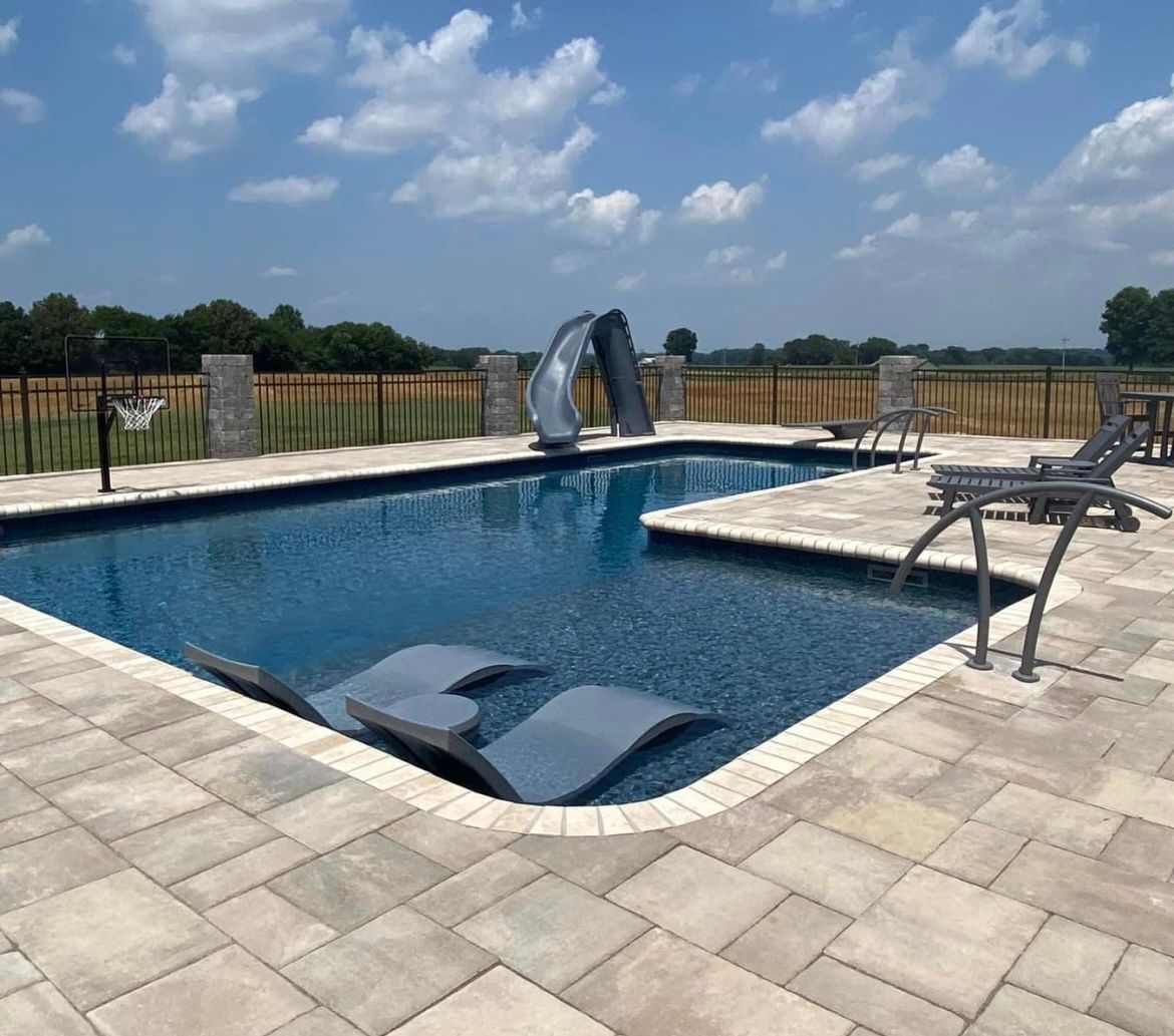 A rectangular swimming pool with a slide, lounge chairs, and a paved patio, against a blue sky.