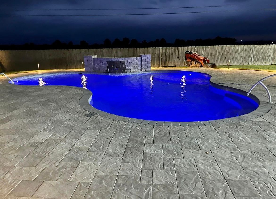 Night view of a swimming pool with blue lights, waterfall feature, and paved patio. Wooden fence in background.