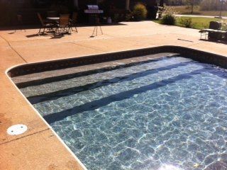 Swimming pool with built-in steps, sparkling blue water, surrounded by concrete patio.