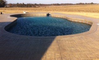 A kidney-shaped swimming pool with blue water and stone patio, set in a field.
