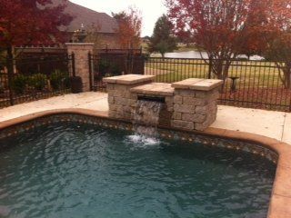 Pool with stone waterfall feature, surrounded by concrete deck, black fence, and trees.