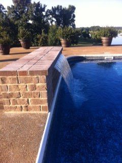 Brick waterfall feature cascading into a blue swimming pool, with planters and trees in the background.