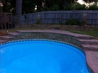 Blue swimming pool with brick steps and a stone wall against a wooden fence.