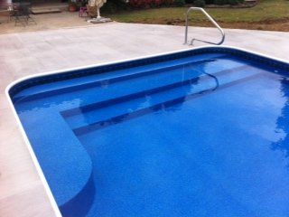 Blue-tiled swimming pool with steps and a metal handrail, surrounded by a concrete patio.